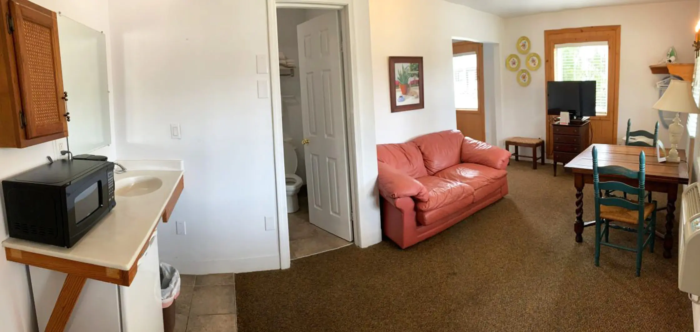 A common room with a red loveseat, a wooden dining table and chairs, and a wall-mounted television, with an open door to a bathroom and a microwave on a counter to one side