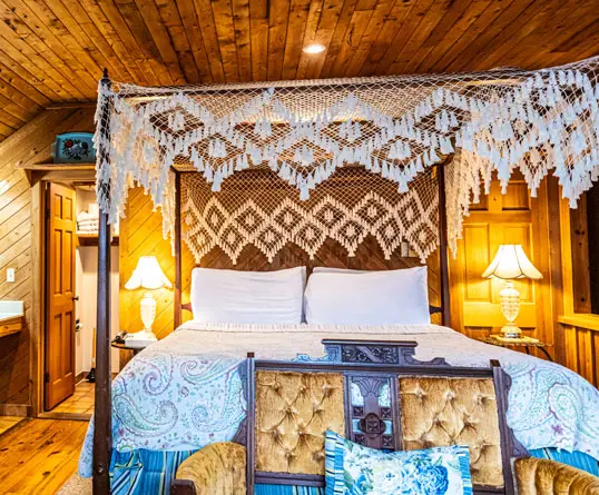 A king bed with an ornate white lace canopy and a patterned quilt, flanked by two lit lamps against a wood-panelled wall