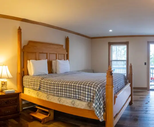 A wooden king bed with a blue plaid quilt in a room with a window and pale wood flooring