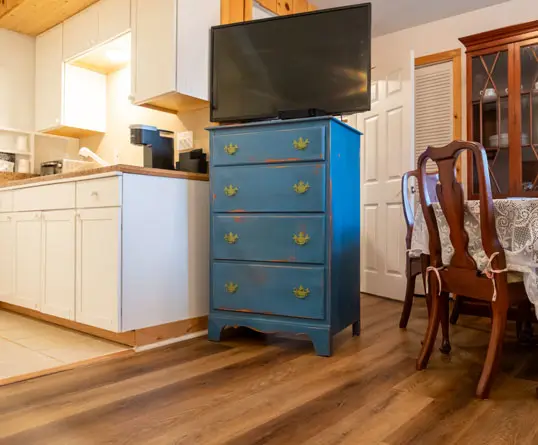 A small white kitchenette with a wall-mounted television above, a blue painted chest of drawers, and dining chairs nearby