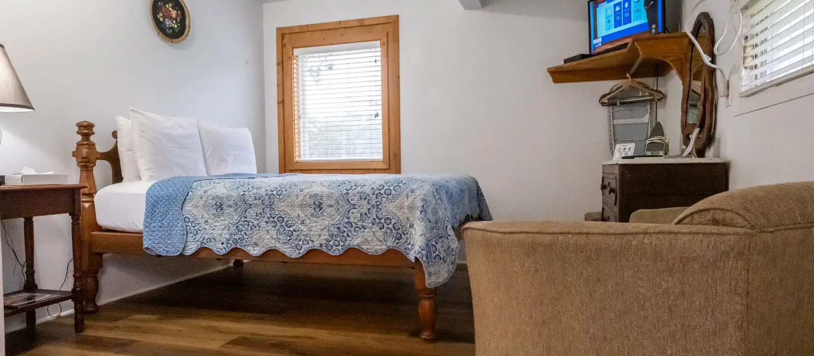 A bed with a blue quilt beside a window, a tan couch, and a small side table with a lantern-style lamp