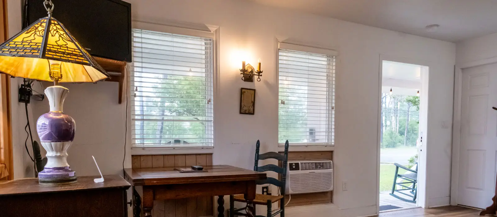 A writing desk with a stained-glass-style lamp beside a window, with a rocking chair visible on the porch outside