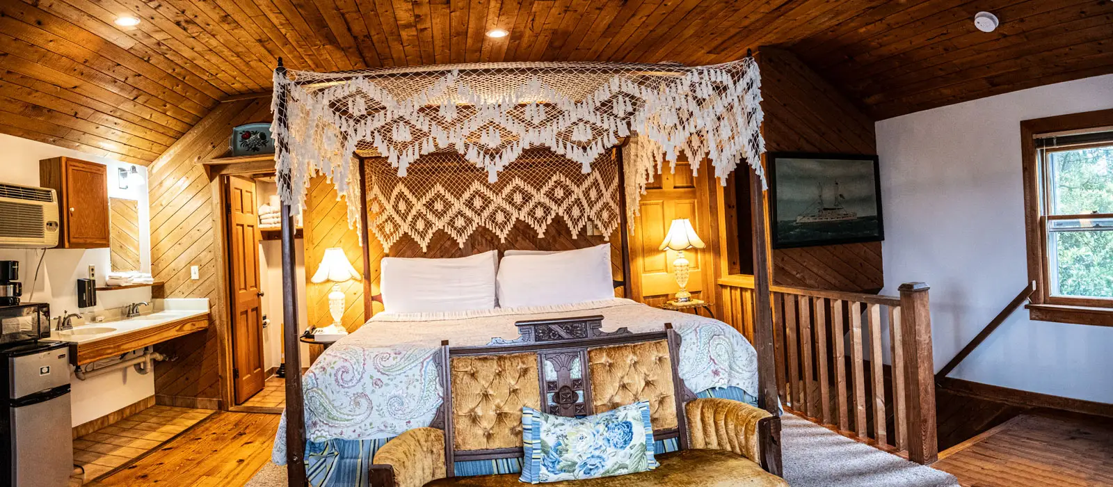 A canopy bed with a patterned quilt beneath a wood-planked ceiling, with a small kitchenette to one side