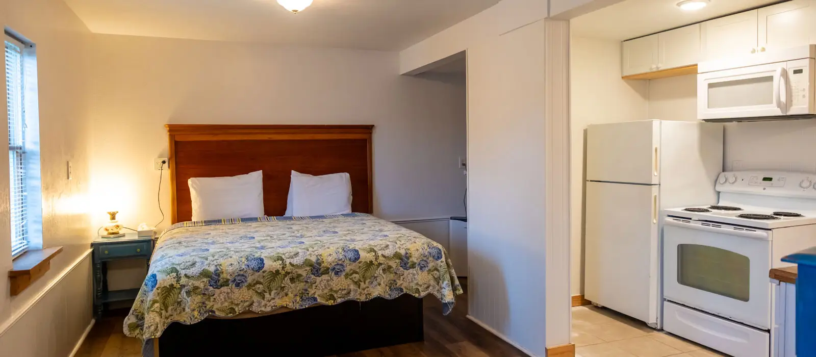 A bed with a floral quilt beside a small kitchenette with a white refrigerator and stove