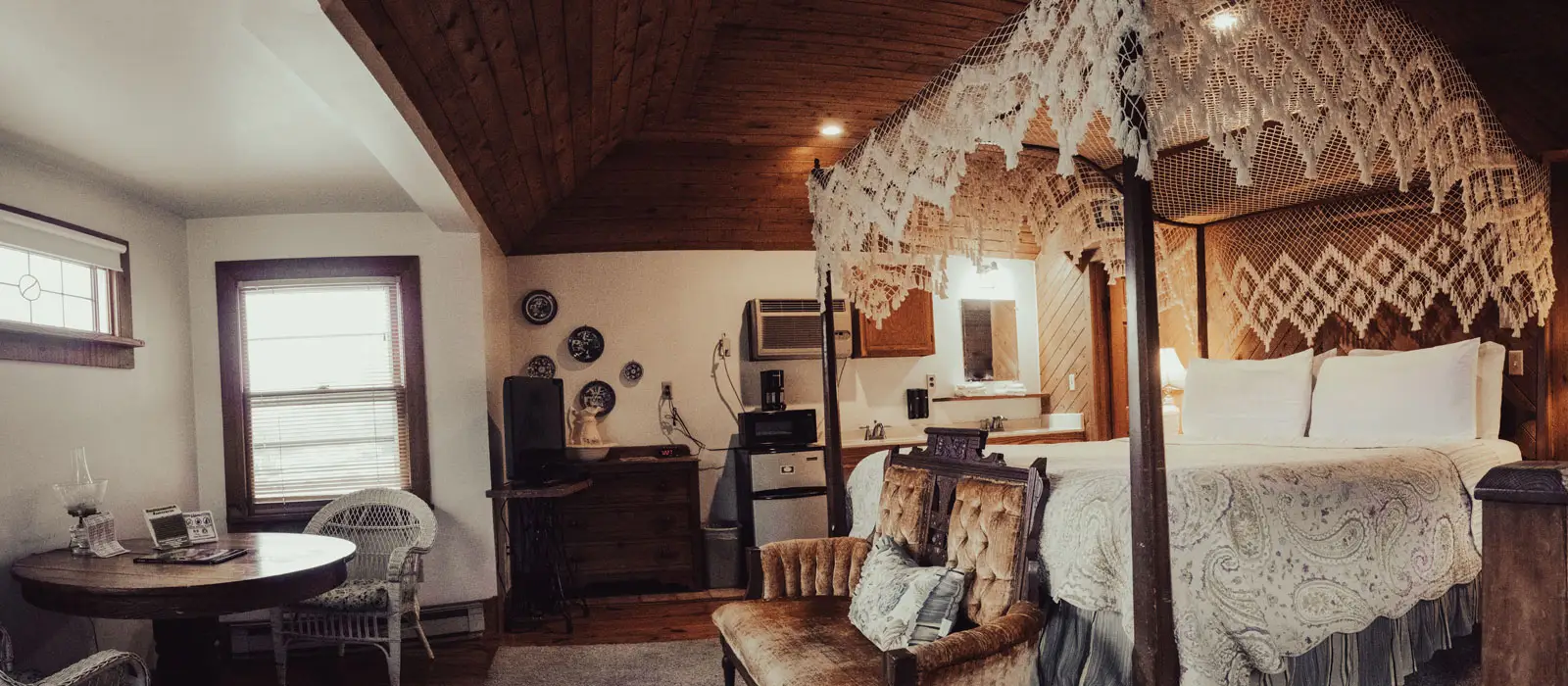 A loft bedroom with a canopy bed, a small round table and chairs, lit warmly under a wood-planked ceiling