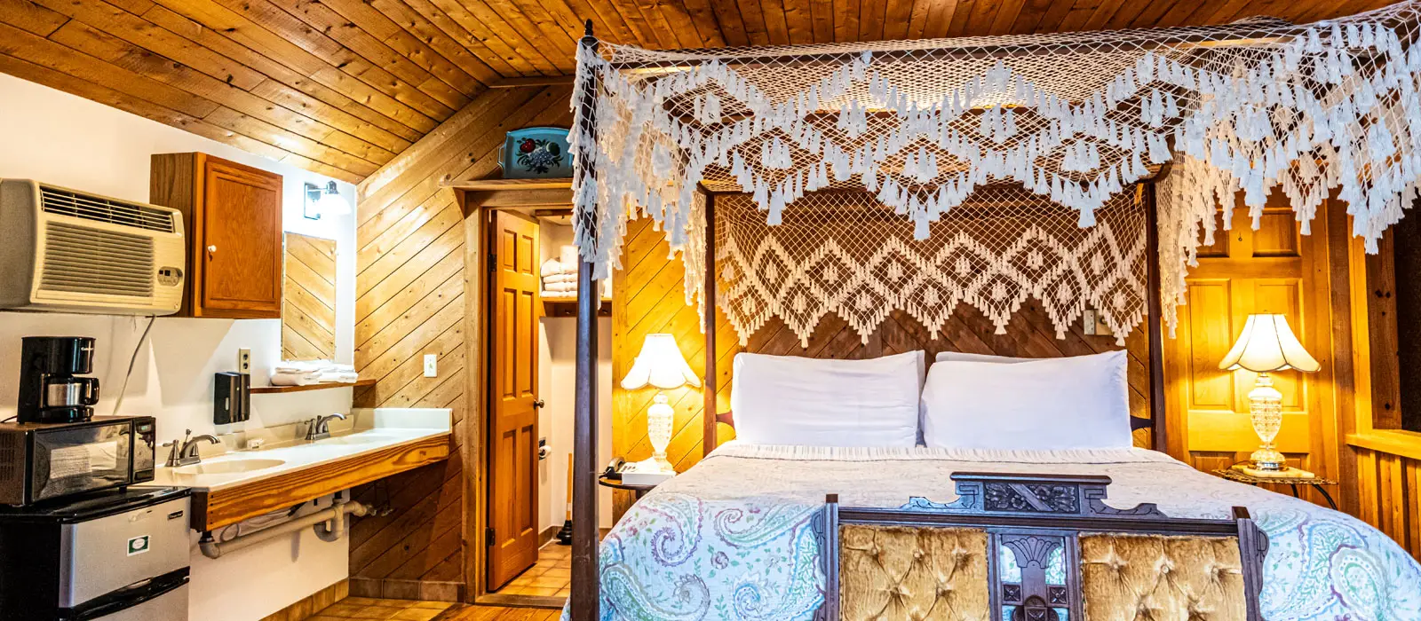 A canopy bed beside a small kitchenette counter with a microwave and coffee maker, under a wood-planked ceiling