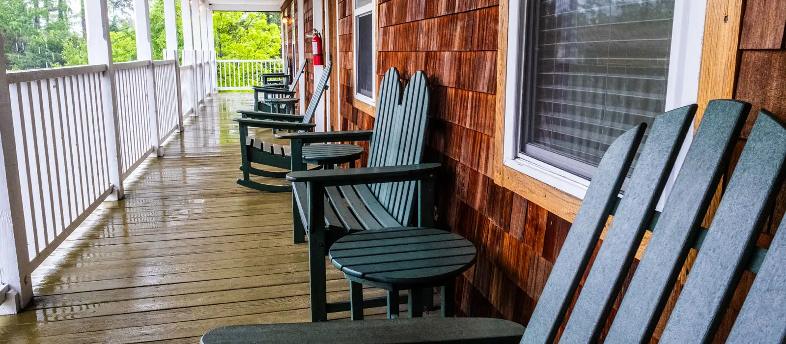A covered porch with green rocking chairs and folded Adirondack chairs, overlooking trees through the railing