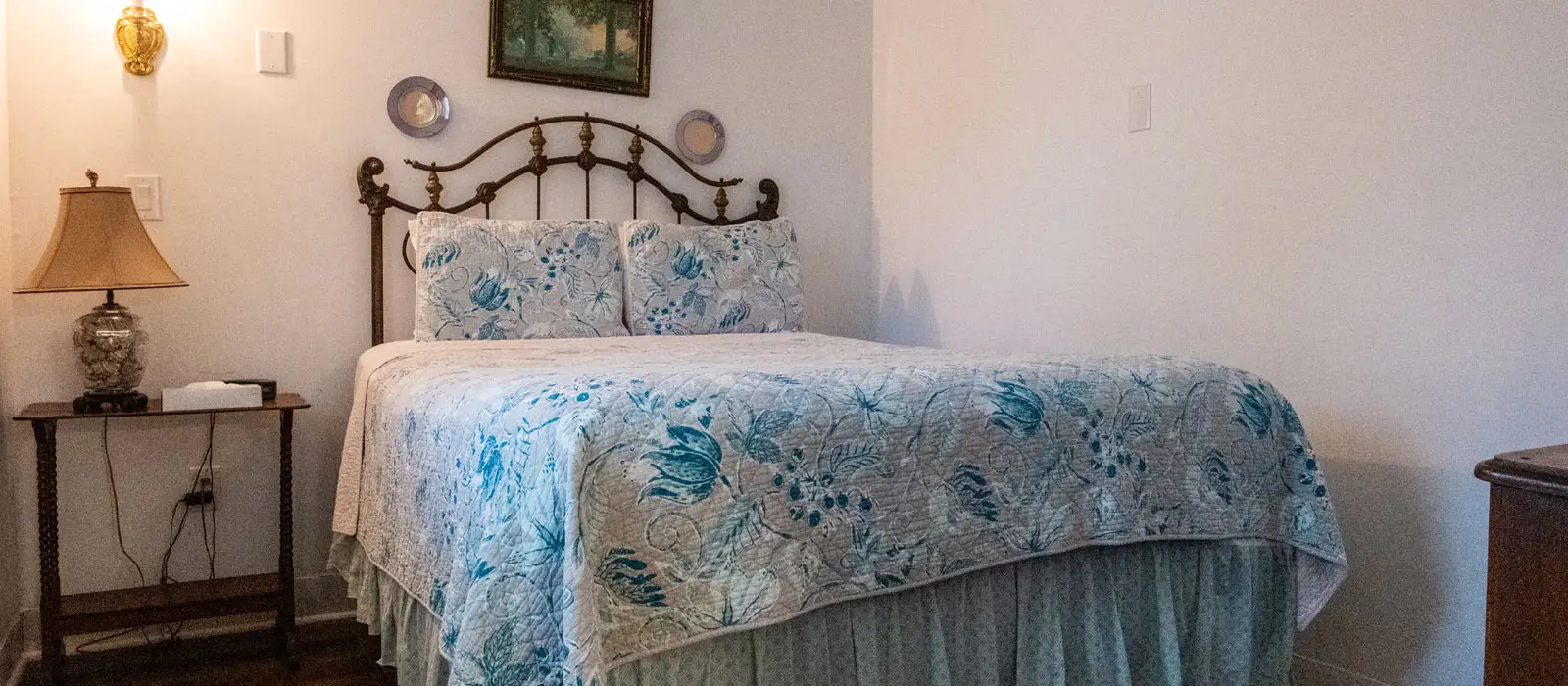 A wrought-iron bed with a blue floral quilt beside a nightstand with a lamp, in a room with patterned wallpaper