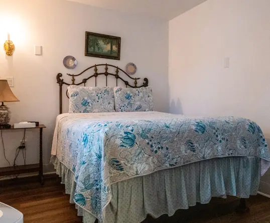 A wrought-iron queen bed with a blue floral quilt against a plain white wall, with a small wall lamp on the nightstand