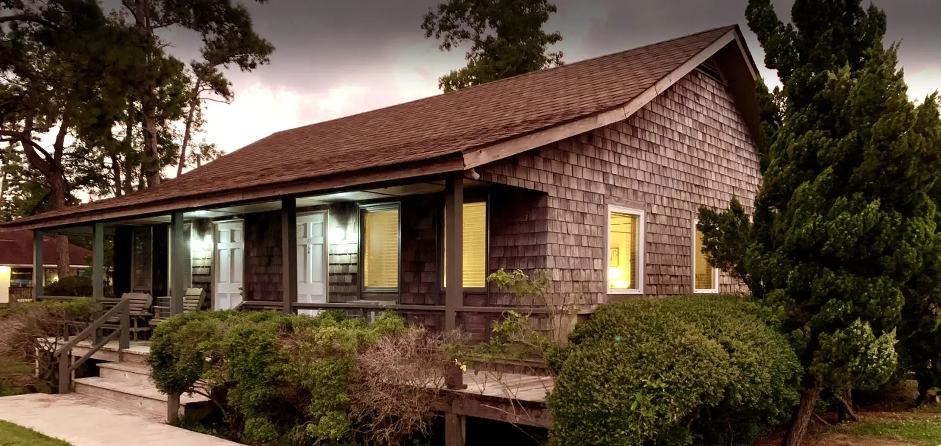 A single-storey shingled building with a covered porch and warmly lit windows, photographed at dusk