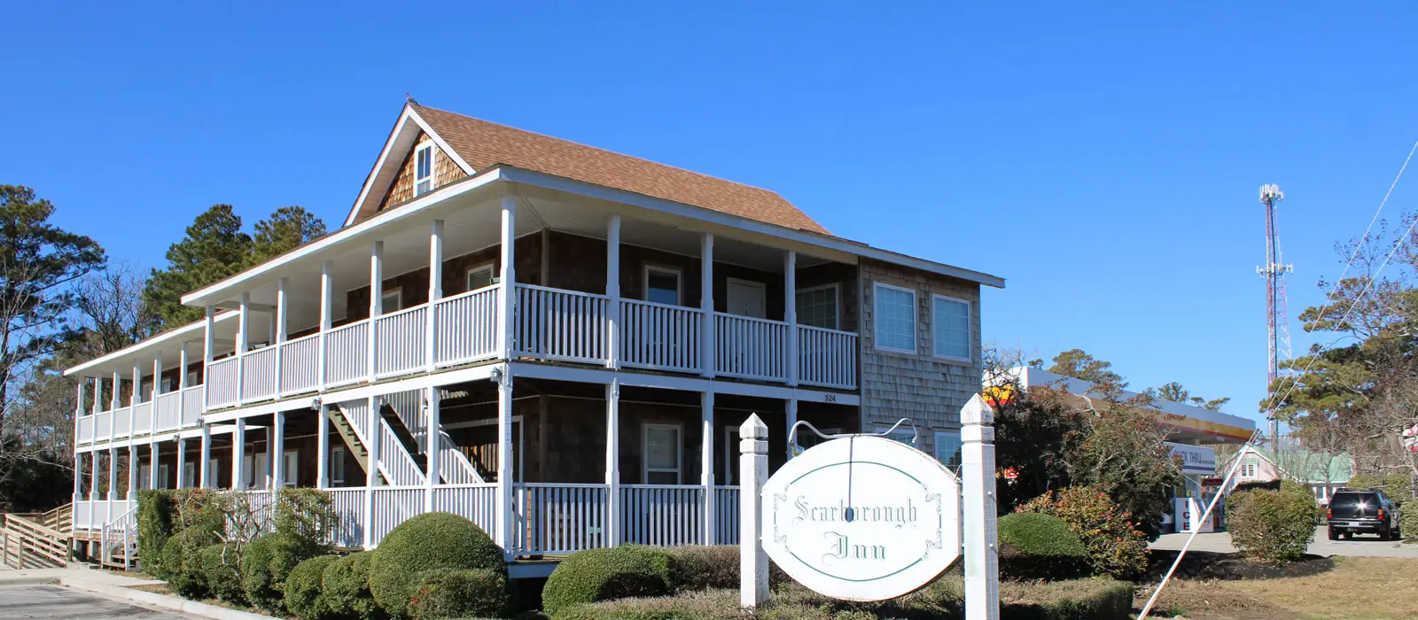 A two-storey shingled building with a wraparound porch and the inn’s oval sign in front, under a clear blue sky