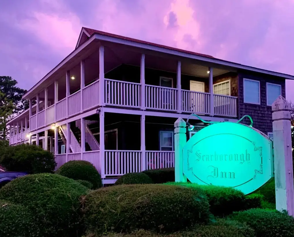 The Scarborough Inn's building at dusk under a purple sky, with its oval sign lit up in front of the porch