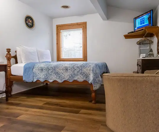 A wooden queen bed with a white and blue quilt beneath a window, with a wall-mounted television in the corner